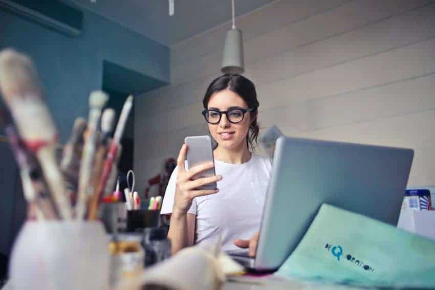 Woman working on phone and laptop at desk with paintbrushes