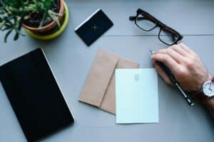 A hand holding a pencil over a desk with a notepad, envelope, and tablet, prepared to do business writing.