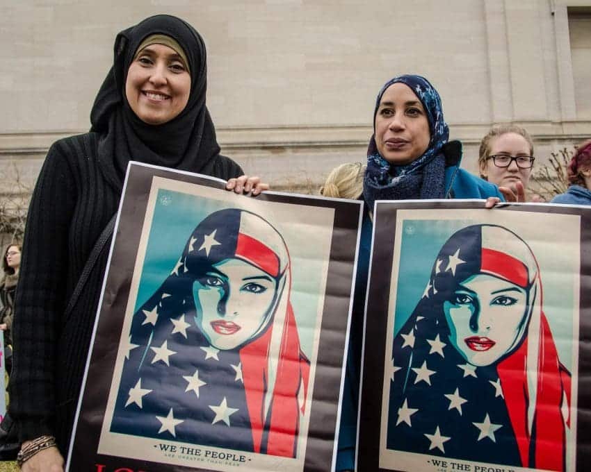 Two Muslim women holding up signs of woman in hijab above “We The People”.