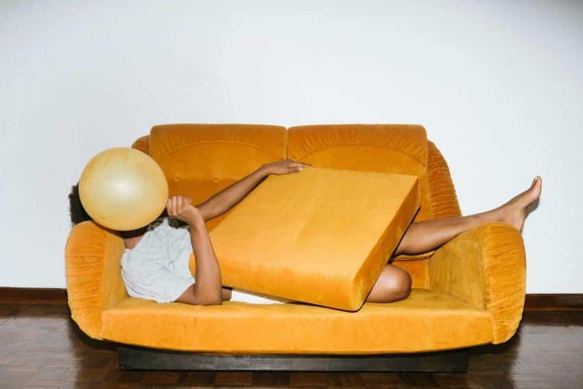 An introverted man lying on a yellow sofa covering his face with a balloon