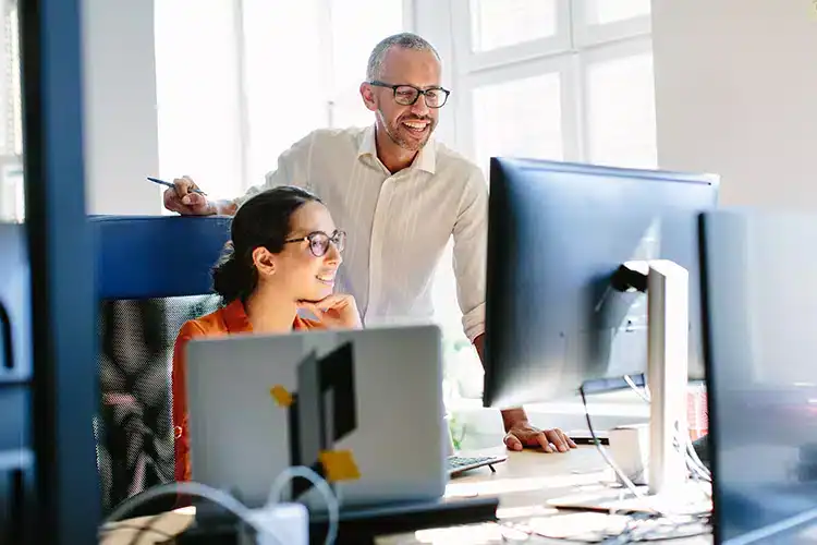 a senior male IT professional guiding a junior female coworker seated in front of her computer