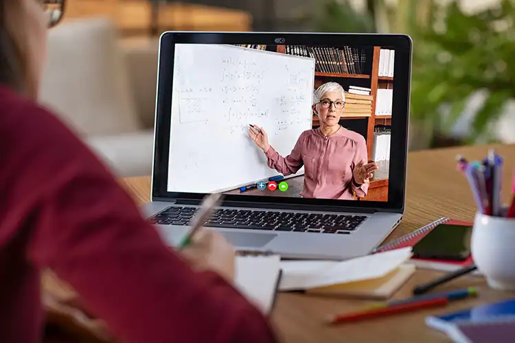 a laptop screen on the desk of an online college student, featuring a female computer science professor teaching a class