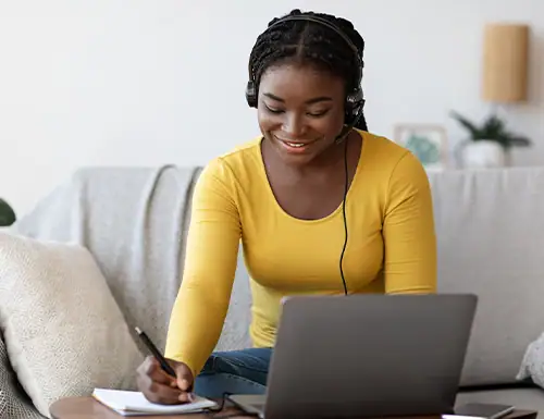 Young woman with headphones using a laptop and writing notes at home, engaging in online education or remote learning.