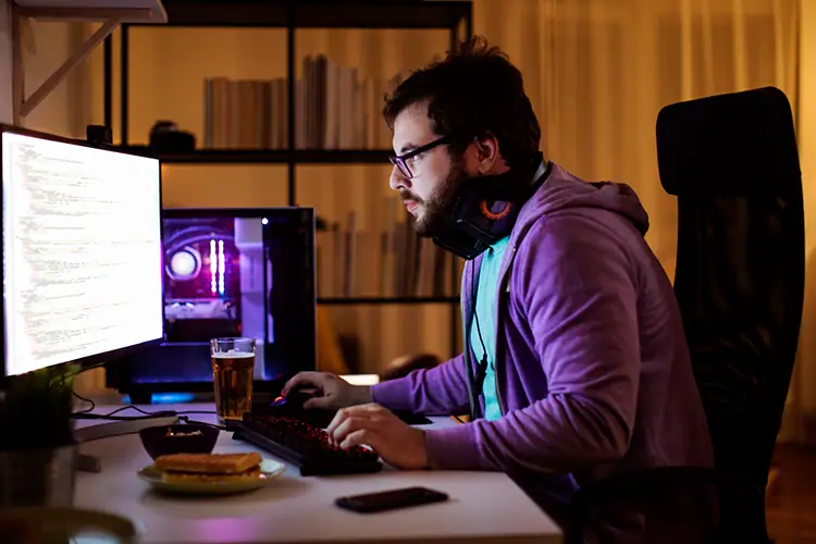 Young man programming at his desk with dual monitors, wearing headphones, in a dimly lit room, focused on game design work.