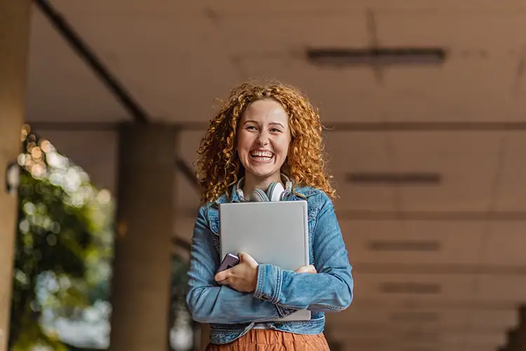 Happy female graduate in cap and gown holding diploma and laptop outdoors, celebrating academic achievement.