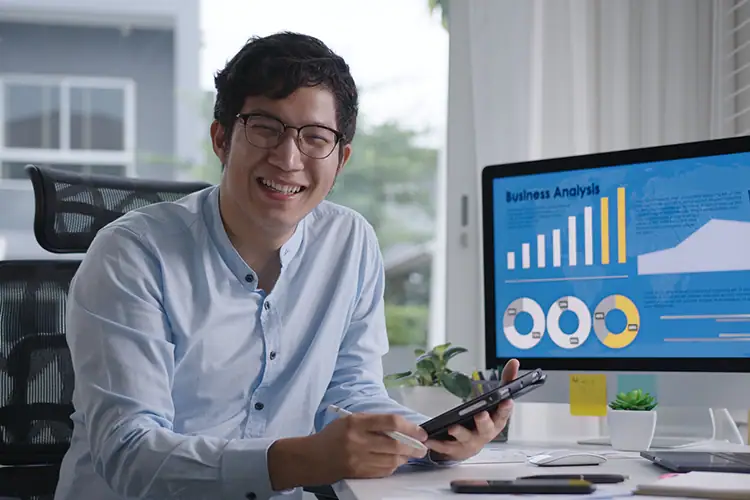 Young man with glasses smiling while analyzing business data on a tablet and computer screen in an office setting.