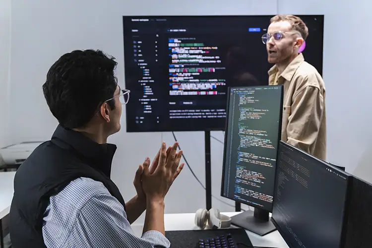 Two developers in a tech office discussing code with multiple monitors showing programming languages and code snippets.