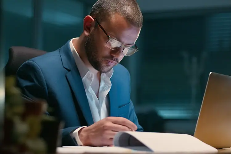A focused professional man in a blue suit and glasses works at a desk with a laptop and documents, studying late at night.