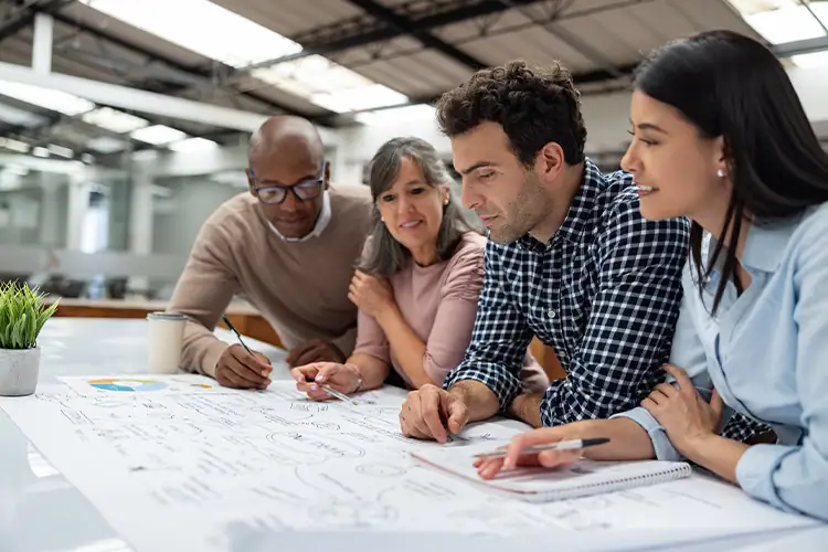 Group of diverse adults collaborating over a large table with documents and notebooks in a modern workspace, focusing on education and teamwork.