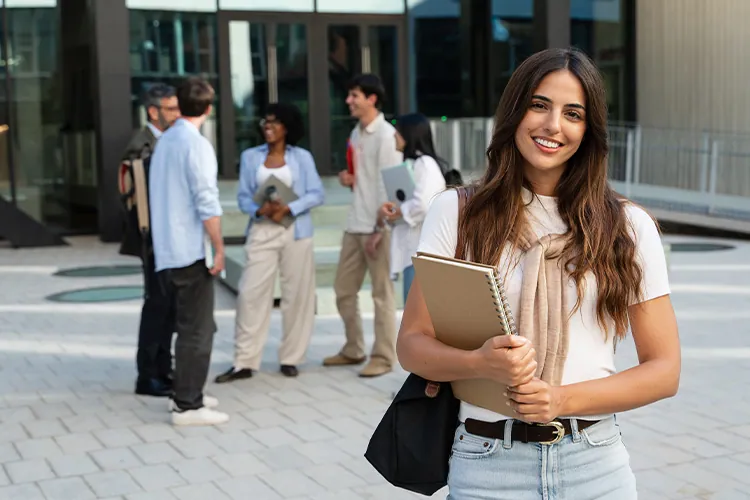 Smiling university student holding notebooks outside a modern campus building with peers in the background.