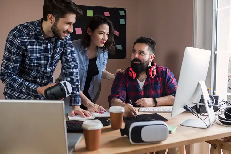 Group of young professionals working together at a modern tech workspace with computers, VR headset, and coffee cups, promoting innovation and teamwork.
