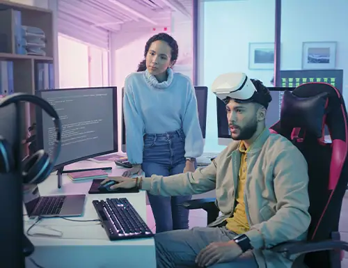 Young man using virtual reality headset with a woman observing in a modern tech lab setting, emphasizing innovation in computer science education.