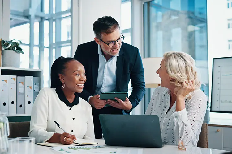 Professional office scene with three colleagues collaborating around a laptop and tablet in bright modern workspace.