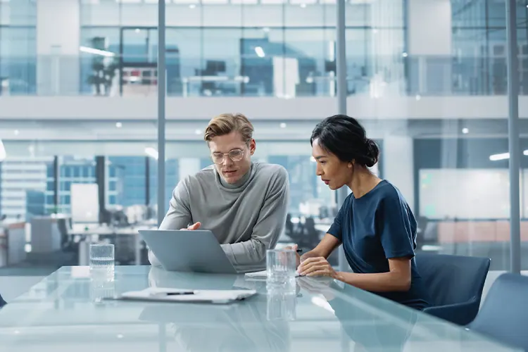 Two professionals discuss data and a laptop at a glass conference table in a modern office setting, representing corporate finance teamwork.